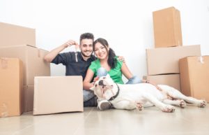 Smiling couple sitting on the floor surrounded by boxes holding the key to their home. A white dog lays at their feet